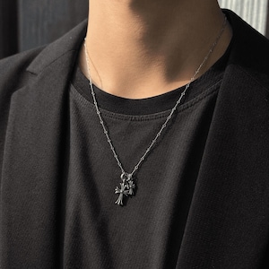 May include: A silver-colored necklace with a double cross pendant. The necklace is worn against a black t-shirt and a black blazer. The cross pendant has intricate detailing.