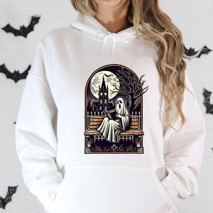 Op de afbeelding: Witte hoodie met een afbeelding van een geest die op een bankje zit voor een spookhuis. De afbeelding is in zwart-wit en heeft een vintage look. De hoodie heeft een capuchon met trekkoord en een kangoeroezak.