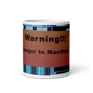 May include: White ceramic mug with a red and orange warning sign that reads "Warning!! Danger to Manifold". The background is a blurry image of a control panel with blue and green lights.