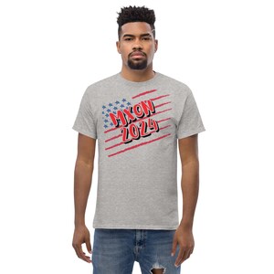 May include: A light gray t-shirt featuring the text "NIXON 2024" in red and blue, with a stylized American flag design. The shirt is a classic crew neck style.