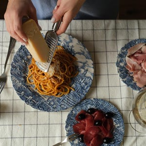Può includere: Una persona sta grattugiando formaggio su un piatto di pasta con un motivo blu e bianco. Ci sono tre piatti su una tovaglia bianca a scacchi. Un piatto ha una fetta di prosciutto, un altro ha una fetta di carne e il terzo ha pasta. C'è un bicchiere di vino bianco sul tavolo.