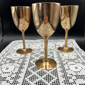 Puede incluir: Tres copas de vino de metal de color dorado con diseño de tallo. Las copas están dispuestas en forma triangular sobre un mantel de encaje blanco.