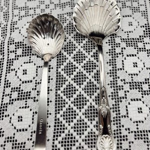 Puede incluir: Dos cucharas de servir de plata con forma de concha. La cuchara más grande tiene un mango decorativo con un diseño de concha. La cuchara más pequeña tiene un cuenco perforado y la palabra "Gianetta" grabada en el mango.