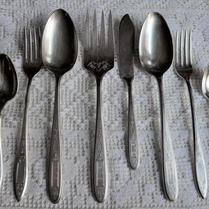 Può includere: Un assortimento di posate d'epoca, tra cui cucchiai, forchette e un coltello da burro, disposte su una tovaglia di pizzo bianco. Le posate presentano dettagli ornati sui manici e una superficie lucida e riflettente.