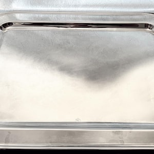 Puede incluir: Una bandeja rectangular plateada con esquinas redondeadas y un borde elevado. La bandeja tiene una superficie pulida y reflectante, y es adecuada para servir alimentos o exhibir artículos. La bandeja mide aproximadamente 46 cm de largo y 30 cm de ancho.