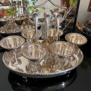 May include: A silver egg cup set with six egg cups, spoons, and a serving tray. The egg cups have a pedestal base and a decorative rim. The spoons are slender and have a rounded bowl. The tray has a beaded edge and a handle.
