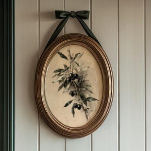 May include: Oval framed botanical print featuring a branch with dark berries and green leaves. The print is in a gold-colored frame and is suspended by a dark green ribbon tied in a bow. The artwork is mounted on a white paneled wall.