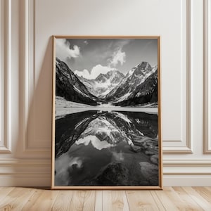 May include: Black and white framed print of a mountain landscape reflected in a lake. The image features snow-capped peaks, a calm lake, and a cloudy sky. The frame is a light brown color, and the print is displayed against a white wall and wooden floor.