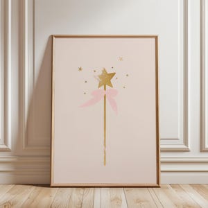 May include: A framed print featuring a whimsical illustration of a gold star-topped wand with a pink bow. The wand is set against a soft pink background, with small gold stars scattered around it. The artwork is framed in a light wood.