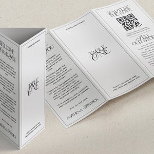 May include: White wedding table cards with black text. One card is folded into a triangular shape, displaying "Welcome, Thank You" and other text. Another card is open, revealing "Table One" and a QR code. The cards are for Athena and Patrick.