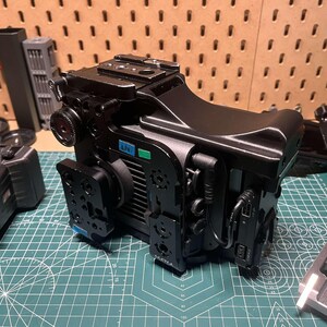 Può includere: Una fotocamera nera con una varietà di accessori, tra cui una gabbia, una maniglia superiore e una maniglia laterale. La fotocamera è appoggiata su un tappetino da taglio verde.