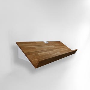 Puede incluir: Estante de madera en ángulo con acabado de vetas naturales. Montado sobre un soporte blanco, creando un efecto flotante. El diseño es minimalista y moderno, adecuado para exhibir libros o decoración.