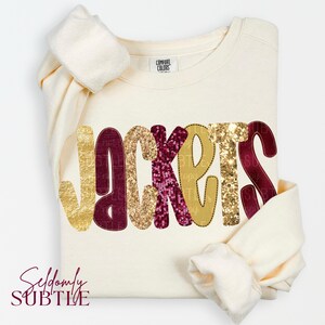 May include: A cream-colored long-sleeve sweatshirt featuring the word JACKETS across the front. The letters are decorated with alternating gold and maroon sequins, and the inner neck label displays the Comfort Colors brand name.