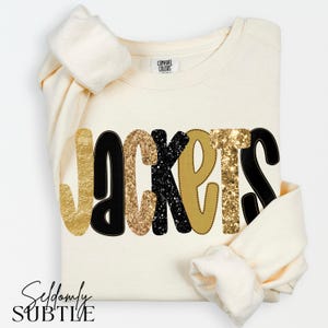 May include: A cream-colored crewneck sweatshirt featuring the word JACKETS printed across the front. The letters alternate between solid black, gold glitter, and matte gold textures. The inner neck label displays the text COMFORT COLORS.