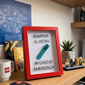 May include: A red framed sign with the text "ROMPERE IL VETRO IN CASO DI EMERGENZA" and a teal Lego remote control. The sign is on a wooden shelf with Lego sets, a mug, and a small succulent.