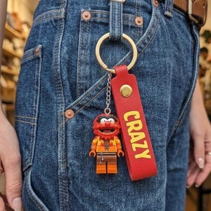 May include: A red leather keychain with the word "CRAZY" in yellow lettering, attached to a gold ring and a small toy figure of Animal from The Muppets. The keychain is attached to a pair of blue denim jeans.