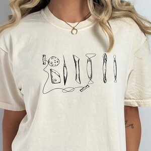 May include: A woman wearing a cream colored t-shirt with a black graphic of various tools. The graphic includes a small circle, a long thin rectangle, a short rectangle, a curved rectangle, a small rectangle, and a long thin rectangle. The woman is also wearing black biker shorts.