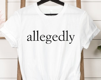 May include: White t-shirt with the word "allegedly" printed in black. The shirt is hanging on a black hanger against a white wood-paneled wall. A wooden ladder and green plant are in the background.