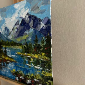 May include: An oil painting of a mountain landscape with a lake. The artwork features a blue sky with white clouds, dark blue mountains, and green trees. The lake reflects the sky and mountains.