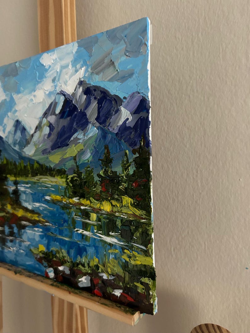 May include: An oil painting of a mountain landscape with a lake. The artwork features a blue sky with white clouds, dark blue mountains, and green trees. The lake reflects the sky and mountains.