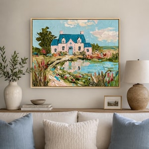 May include: An oil painting in a gold frame depicting a pink house with a blue roof situated beside a reflective pond. The scene includes thick, textured brushstrokes, green trees, and colorful wildflowers under a light blue sky with clouds.
