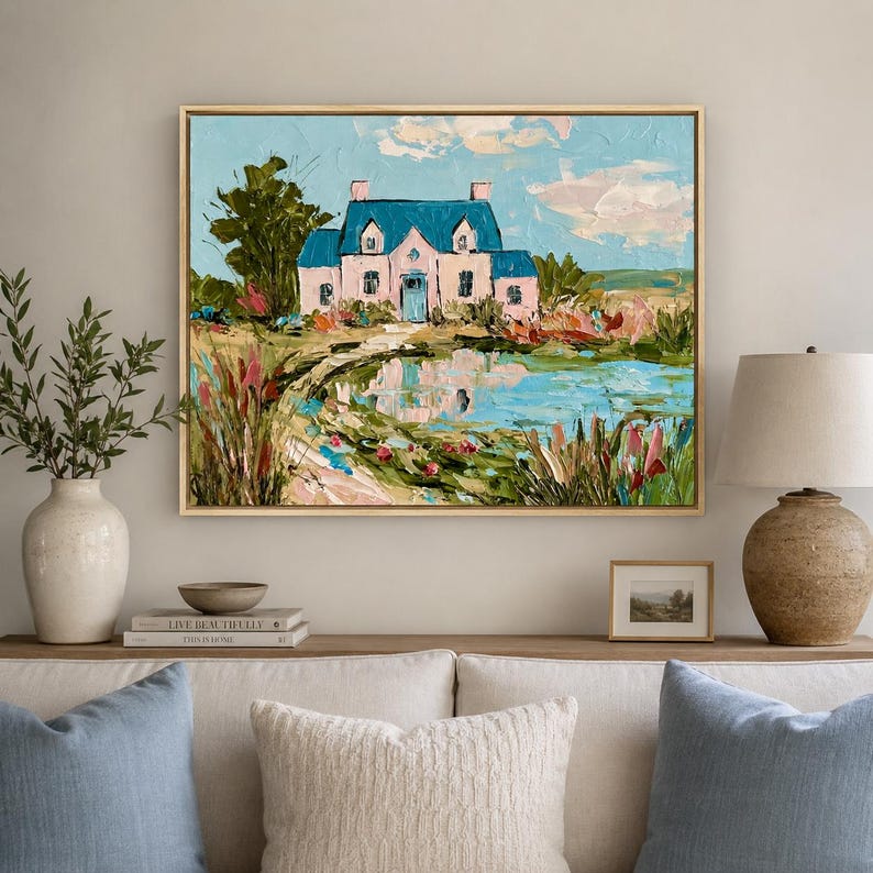May include: An oil painting in a gold frame depicting a pink house with a blue roof situated beside a reflective pond. The scene includes thick, textured brushstrokes, green trees, and colorful wildflowers under a light blue sky with clouds.