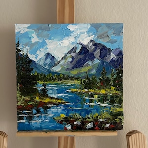 May include: Oil painting of a mountain landscape with a lake. The artwork features a blue lake reflecting the sky and mountains. The mountains are shades of blue and green, with green trees along the lake's edge. The sky is a mix of blue and white.
