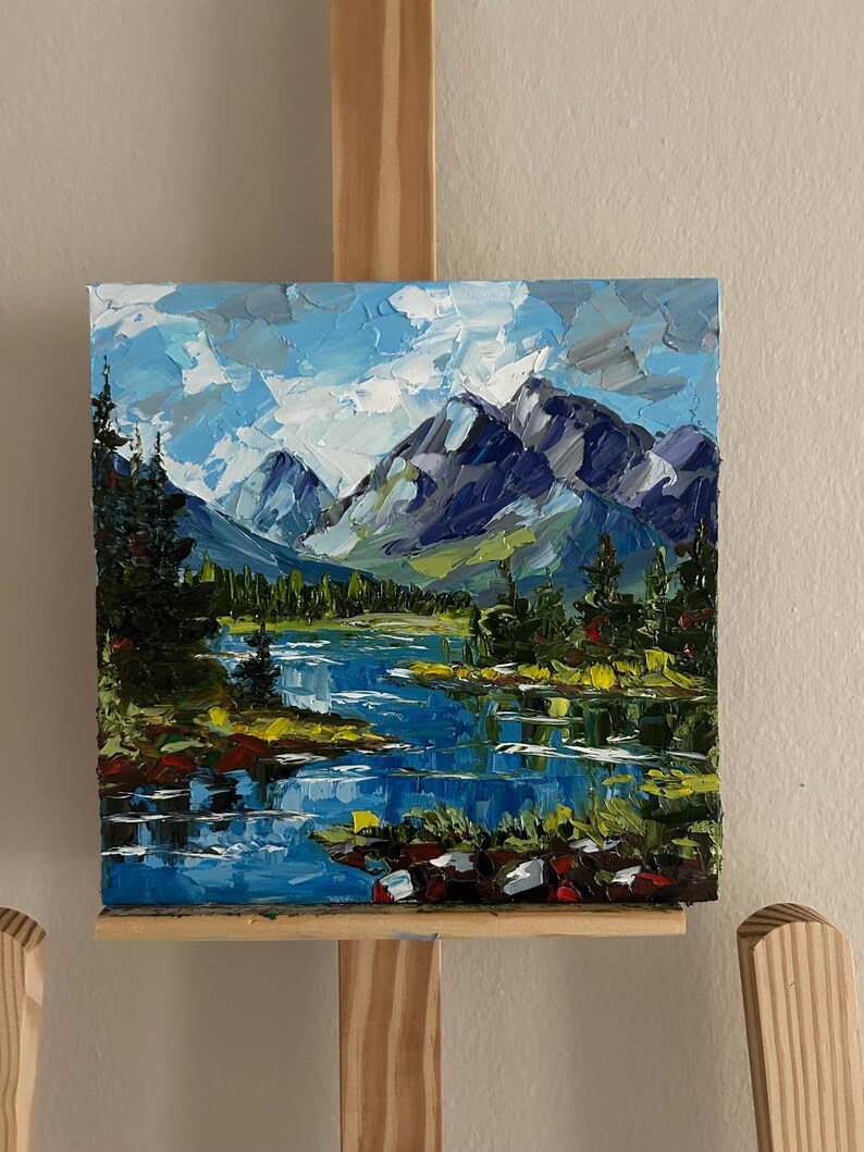 May include: Oil painting of a mountain landscape with a lake. The artwork features a blue lake reflecting the sky and mountains. The mountains are shades of blue and green, with green trees along the lake's edge. The sky is a mix of blue and white.
