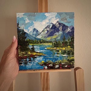 May include: An oil painting of a mountain landscape with a blue lake and evergreen trees. The sky is a mix of blue and white, and the mountains are shades of gray and purple. The painting is displayed on a wooden easel.