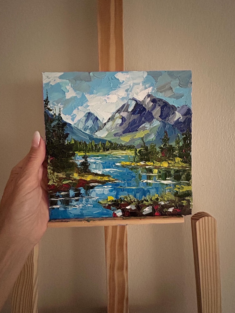 May include: An oil painting of a mountain landscape with a blue lake and evergreen trees. The sky is a mix of blue and white, and the mountains are shades of gray and purple. The painting is displayed on a wooden easel.