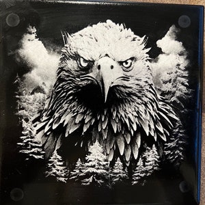 Könnte beinhalten: Quadratischer schwarzer Untersetzer mit einer detaillierten Graustufenillustration eines Adlerkopfes. Weiße Kiefern und Wolken bilden den Hintergrund und erzeugen einen auffälligen Kontrast. Der Adler hat einen intensiven Blick.