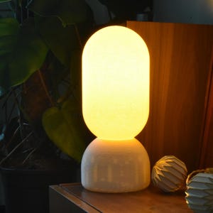 Puede incluir: Una l&aacute;mpara de mesa de cer&aacute;mica blanca con una pantalla redonda de vidrio esmerilado que emite una luz c&aacute;lida de color amarillo. La l&aacute;mpara est&aacute; colocada sobre una superficie de madera junto a un objeto decorativo.