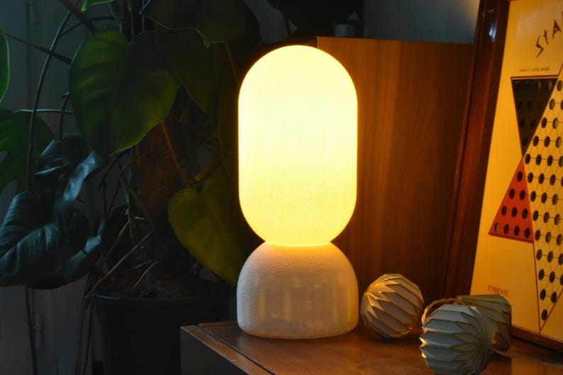 Puede incluir: Una l&aacute;mpara de mesa de cer&aacute;mica blanca con una pantalla redonda de vidrio esmerilado que emite una luz c&aacute;lida de color amarillo. La l&aacute;mpara est&aacute; colocada sobre una superficie de madera junto a un objeto decorativo.