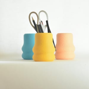 Peut inclure: Trois organiseurs de bureau de couleur bleue, jaune et pêche. Les organiseurs sont faits d'un matériau texturé et ont une forme arrondie. Une paire de ciseaux et des crayons sont dans l'organisateur jaune.