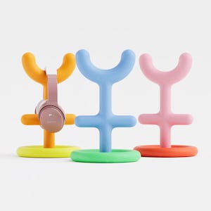 May include: Three brightly coloured headphone stands in yellow, blue, and pink. One stand holds rose-gold headphones. Each stand has a circular base and a Y-shaped top, designed to hold headphones.
