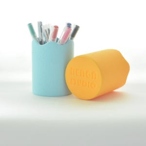 Peut inclure: Un pot à crayons bleu clair avec un bord festonné, contenant des stylos de différentes couleurs. À côté, un objet cylindrique orange avec le texte "RENGU STUDIO". Les objets sont sur une surface blanche.