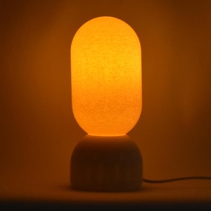 Peut inclure: Une lampe de table blanche avec une base ronde, texturée et blanche et un abat-jour rond, texturé et blanc. L'abat-jour est éclairé de l'intérieur, projetant une lueur orange chaude.