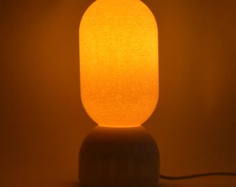 Puede incluir: Una lámpara de mesa blanca con una base redonda, texturizada y blanca y una pantalla redonda, texturizada y blanca. La pantalla está iluminada desde el interior, proyectando un cálido resplandor naranja.