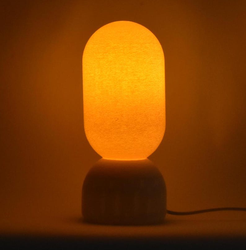 Puede incluir: Una l&aacute;mpara de mesa blanca con una base redonda, texturizada y blanca y una pantalla redonda, texturizada y blanca. La pantalla est&aacute; iluminada desde el interior, proyectando un c&aacute;lido resplandor naranja.