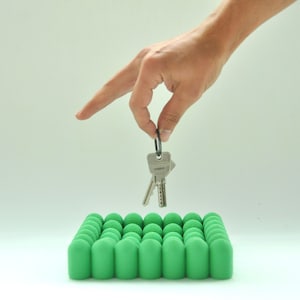 Op de afbeelding: Een set sleutels hangt boven een groene, getextureerde sleutelhouder. De sleutelhouder is gemaakt van vele kleine, afgeronde cilinders. De sleutels worden vastgehouden door een zwarte ring. De achtergrond is effen licht van kleur.