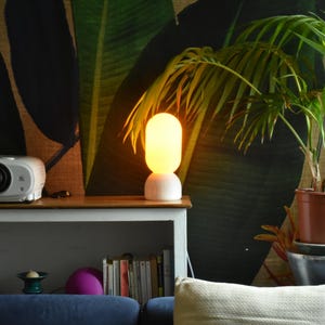 Puede incluir: Una l&aacute;mpara de mesa blanca con una pantalla redonda de vidrio esmerilado que emite una luz c&aacute;lida amarilla. La l&aacute;mpara est&aacute; en un estante de madera con un proyector y libros. Una planta en maceta con hojas verdes est&aacute; en el fondo.
