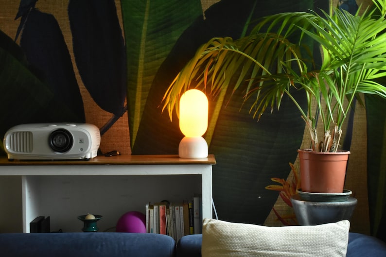Puede incluir: Una l&aacute;mpara de mesa blanca con una pantalla redonda de vidrio esmerilado que emite una luz c&aacute;lida amarilla. La l&aacute;mpara est&aacute; en un estante de madera con un proyector y libros. Una planta en maceta con hojas verdes est&aacute; en el fondo.