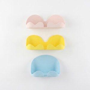 May include: Three wall-mounted storage organizers in pastel colors. The top organizer is light pink, the middle is yellow, and the bottom is light blue. Each organizer features a scalloped front edge and a curved, semi-circular back panel design.