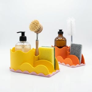 Puede incluir: Un organizador de fregadero amarillo con borde ondulado y base rosa, que contiene un dispensador de jabón transparente, un cepillo de madera y una esponja amarilla. Al lado, un organizador naranja con borde ondulado y base rosa sostiene un dispensador de jabón marrón, un cepillo blanco y una esponja gris.
