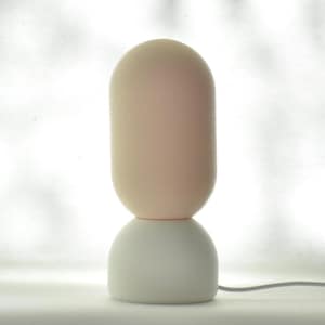 Puede incluir: Una l&aacute;mpara de mesa moderna con una base blanca y una pantalla redonda de color rosa claro. La l&aacute;mpara tiene un dise&ntilde;o simple y minimalista.