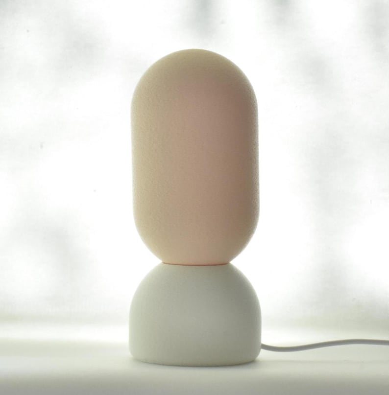 Puede incluir: Una l&aacute;mpara de mesa moderna con una base blanca y una pantalla redonda de color rosa claro. La l&aacute;mpara tiene un dise&ntilde;o simple y minimalista.