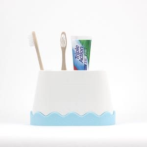 Peut inclure: Un porte-brosse à dents blanc avec une base ondulée bleu clair, contenant deux brosses à dents et un tube de dentifrice. Le tube de dentifrice est vert, bleu et blanc avec le texte "Triple Action".