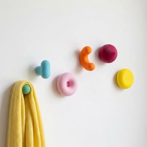 Peut inclure: Un ensemble de cinq patères murales de formes et de couleurs variées, incluant une forme en T bleu clair, un beignet rose, une forme incurvée orange, une sphère rose foncé et un cercle jaune, avec un tissu jaune suspendu à la patère bleue.
