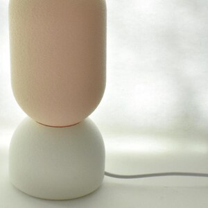Puede incluir: Una l&aacute;mpara de mesa de cer&aacute;mica blanca y rosa con una base redonda y texturizada y un cable.