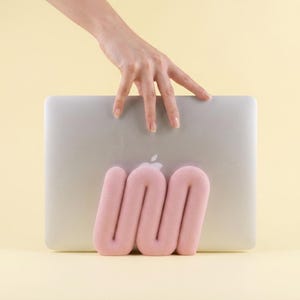 Puede incluir: Un ordenador portátil plateado sostenido en posición vertical por un soporte de espuma suave, de color rosa claro y con forma de serpentina. El soporte presenta un diseño tubular curvo y continuo que sostiene la base del portátil.